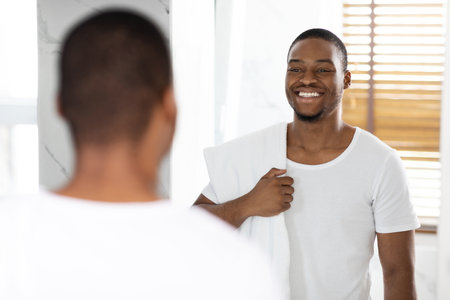 Man Smiling at His Reflection in the Mirror While Holding a Towel in a Bright Bathroomの写真素材