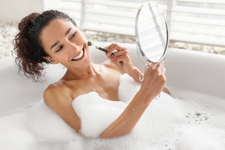 Woman Enjoying a Relaxing Bubble Bath While Applying Skincare in a Bright Bathroom Settingの写真素材