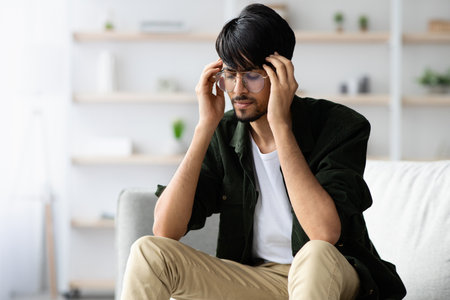 Young Man Sitting on Couch Looking Stressed While Holding His Head in a Bright Living Roomの写真素材