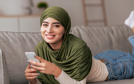 Young Woman in Green Headscarf Smiles While Using Smartphone on a Couch