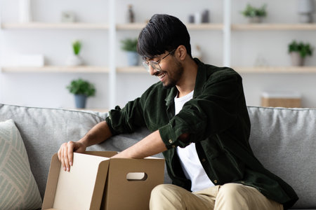 Smiling Man Opens a Cardboard Box While Sitting on a Grey Sofa in a Cozy, Well-Decorated Living Room