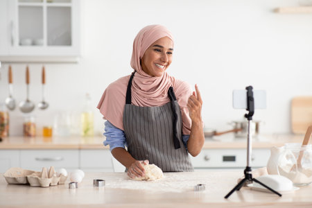 Woman in Apron Demonstrating Dough Preparation in a Bright Kitchen While Interacting With an Online Audienceの写真素材