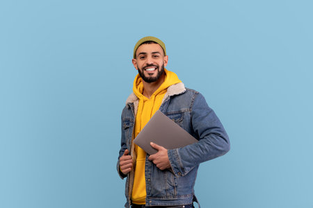 Young Man With Laptop Smiles Against a Blue Background, Showcasing a Casual and Modern Vibe in a Relaxed Settingの写真素材