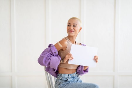 Vaccinated young lady with medical band on shoulder sitting on chair over white wall and showing white empty placardの写真素材