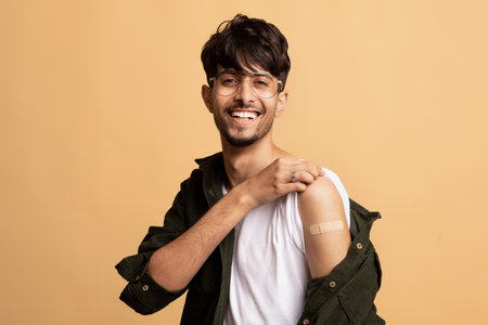 Young Man Smiling Proudly After Getting Vaccinated, Showcasing the Band-Aid on His Arm Against a...