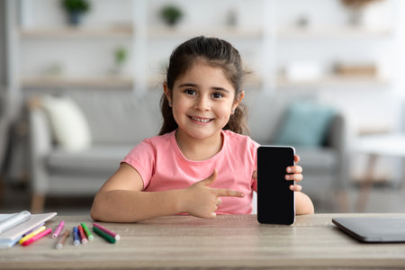 Young Girl Happily Pointing at a Blank Smartphone in a Cozy Indoor Setting While Seated at a Table...