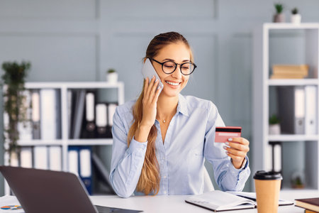 Smiling Woman Making a Phone Call While Holding a Credit Card in Modern Office Setting
