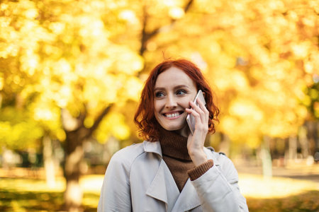 Cheerful Woman Talking on a Smartphone Under Vibrant Autumn Trees in a Sunny Park