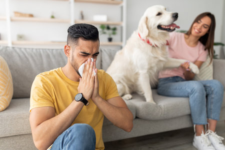 Man Sneezes on Couch While Woman Laughs With Dog in a Cozy Living Room Settingの写真素材