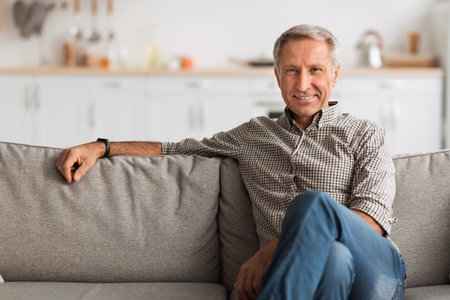 Mature Man Sitting Comfortably on a Couch in a Modern Living Room Looking Relaxed and Cheerfulの写真素材