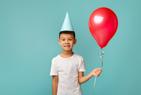 Young Boy Celebrates Birthday Wearing a Party Hat and Holding a Red Balloon Against a Light Blue...