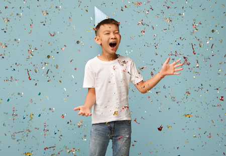 Child Celebrates With Confetti During Birthday Party While Wearing a Party Hat Against a Light Blue Backgroundの写真素材