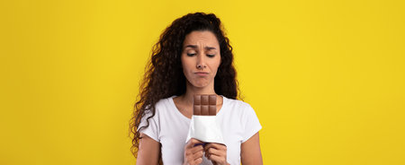 Woman Expresses Disappointment While Holding Chocolate Bar Against Bright Yellow Backgroundの写真素材