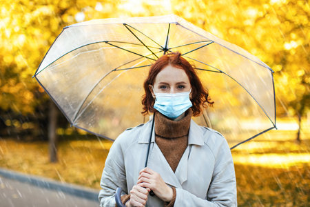 Woman With Red Hair Holding Transparent Umbrella Wearing Mask During Autumn Rain in Urban Parkの写真素材