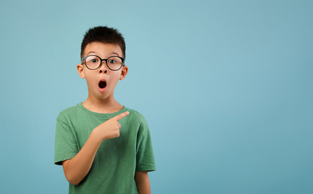 Excited Boy in Green Shirt Expresses Surprise While Pointing to the Side Against a Blue Background