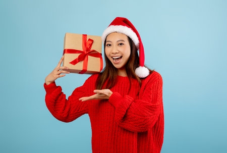 Happy Girl in a Red Sweater and Santa Hat Holding a Christmas Gift Against a Blue Backgroundの写真素材