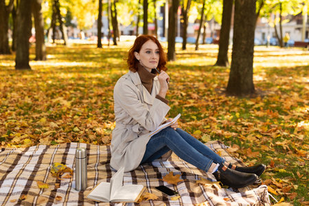 Woman Enjoying a Peaceful Afternoon in a Park Surrounded by Autumn Leaves While Writing in Her Notebookの写真素材