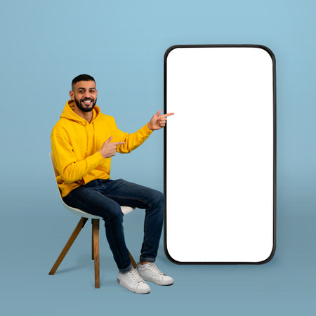 Cheerful Man in Yellow Hoodie Poses Next to Empty Vertical Frame on Blue Background, Inviting...