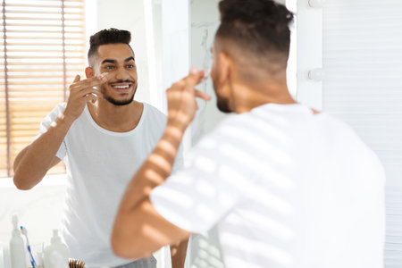 Man Applies Skincare Products While Smiling in a Well-Lit Bathroom in the Morningの写真素材