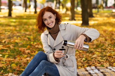 Woman in a Light Coat Pouring Tea Into a Cup While Sitting on a Blanket in a Colorful Autumn Park