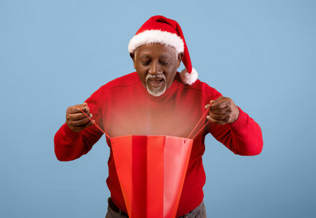 Man in Red Sweater and Santa Hat Opens Gift Bag With Excitement During Holiday Seasonの写真素材