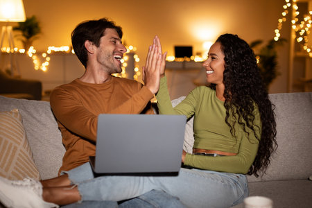 Joyful Couple Celebrates Victory With High-Five While Using Laptop at Home in Cozy Living Roomの写真素材