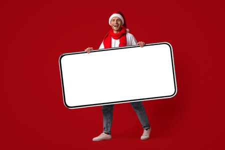 Cheerful Person Wearing a Santa Hat and Scarf Holding a Blank Sign Against a Bright Red Backgroundの写真素材