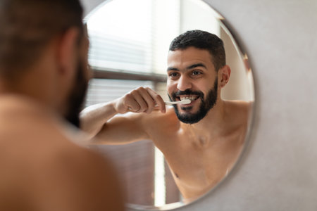 Young Arab Man Brushes Teeth in Modern Bathroom While Smiling at Mirrorの写真素材