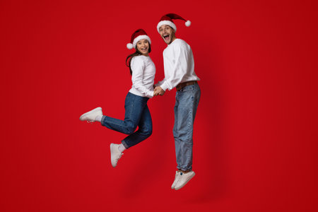 Joyful Couple Celebrating Holidays With Excitement While Jumping Against a Bright Red Backdrop in Festive Attireの写真素材