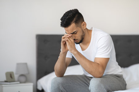 Sad Middle Eastern Man Sitting on Bed Appears Deep in Thought at Homeの写真素材