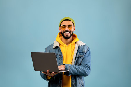 Young Man Smiles While Using Laptop in Casual Outfit Against a Blue Background