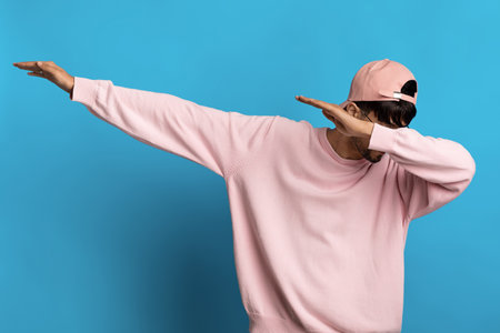 Young Man in a Pink Sweater and Cap Performs a Dab Dance Move Against a Blue Background While Having...