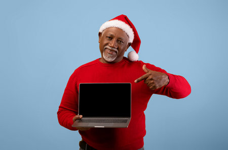 Man in Red Sweater and Santa Hat Points to Laptop Screen for Christmas Celebrationの写真素材