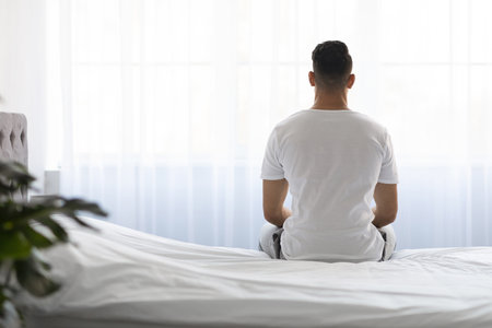 Lonely Young Man Sitting on Bed Looking out Window in Morning Lightの写真素材