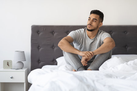 Thoughtful Young Arab Man Sitting on Bed in a Comfortable Room Contemplating Lifeの写真素材
