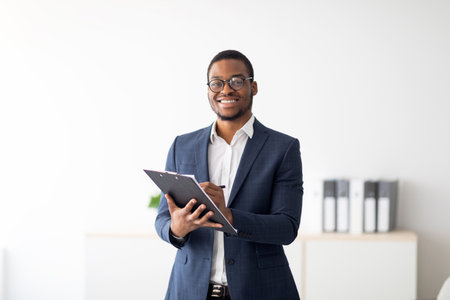 Happy young black male psychologist writing in clipboard, looking at camera and smiling in modern officeの写真素材