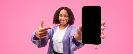 Young Woman Shows Smartphone in Front of Pink Background While Smiling and Pointing at Deviceの写真素材