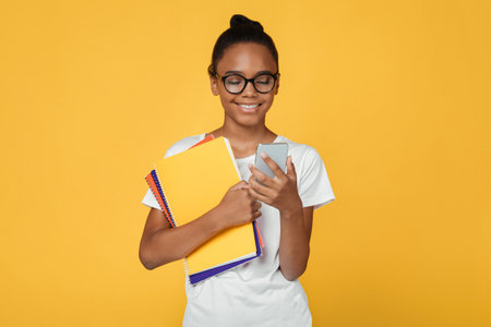 Smiling teenager african american girl pupil in glasses and casual holds books and reads message on...