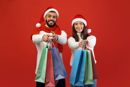 Happy Couple Celebrating Christmas Sales in Santa Hats With Shopping Bags Against Red Backdropの写真素材