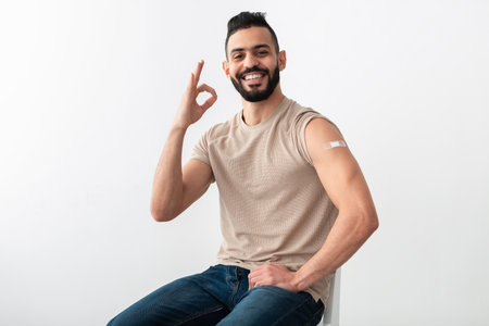 Joyful young Arab guy with vaccinated arm after covid injection showing okay gesture on white studio backgroundの写真素材