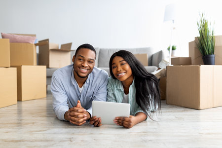 Happy black family lying on floor in new apartment with digital tablet among cardboard boxes with...