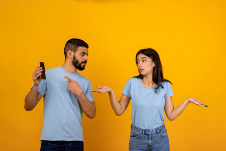 Angry Man Confronts Girlfriend About Provocative Messages on Smartphone Against Yellow Backgroundの写真素材