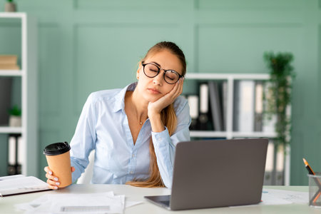 Young Woman in Office Sleeping While Holding Coffee Cup After Long Computer Workの写真素材