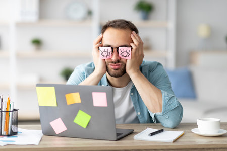 Tired young man with open eyes drawing on his glasses, sleeping in front of laptop at home office