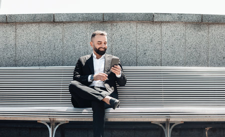 Happy successful mature businessman using smartphone, resting on street, sitting on bench and using mobile applicationの写真素材