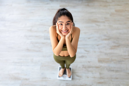 Pretty Indian woman on scales holding her face in excitement, weighing after successful slimming at...