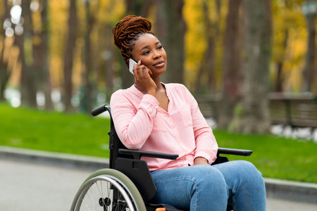 Young Afro lady in wheelchair having phone conversation at city park in autumn