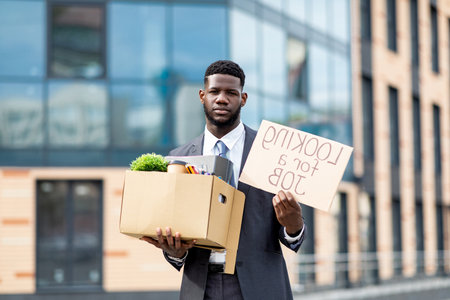 Looking for a job. Upset black businessman with box of personal stuff and poster, got fired, standing outdoorsの写真素材