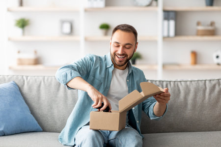 Happy Caucasian man receiving package, unboxing parcel, taking out gaming joystick, sitting on sofa in living roomの写真素材