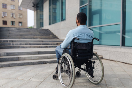 Back view of young black guy in wheelchair standing in front of stairs without ramp, having problem entering buildingの写真素材